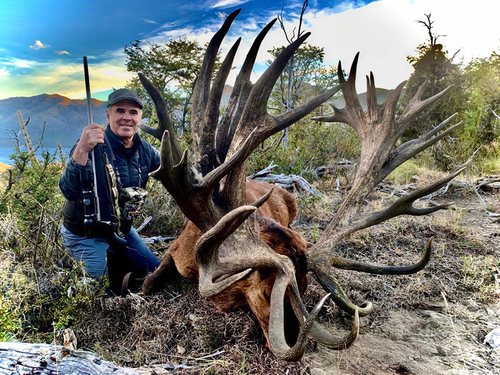Hunter with rifle beside a towering multi-tined red stag above the lake