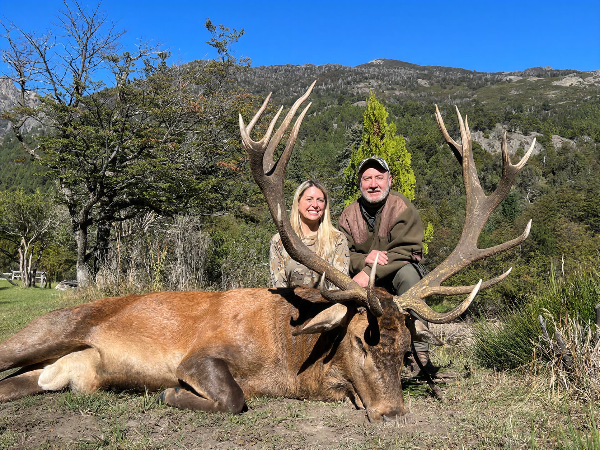 Two hunters with a wide red stag on a sunny Andean meadow
