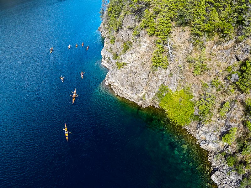 Aerial view of a group of kayakers paddling along a rocky forested shoreline on a deep blue Andean lake