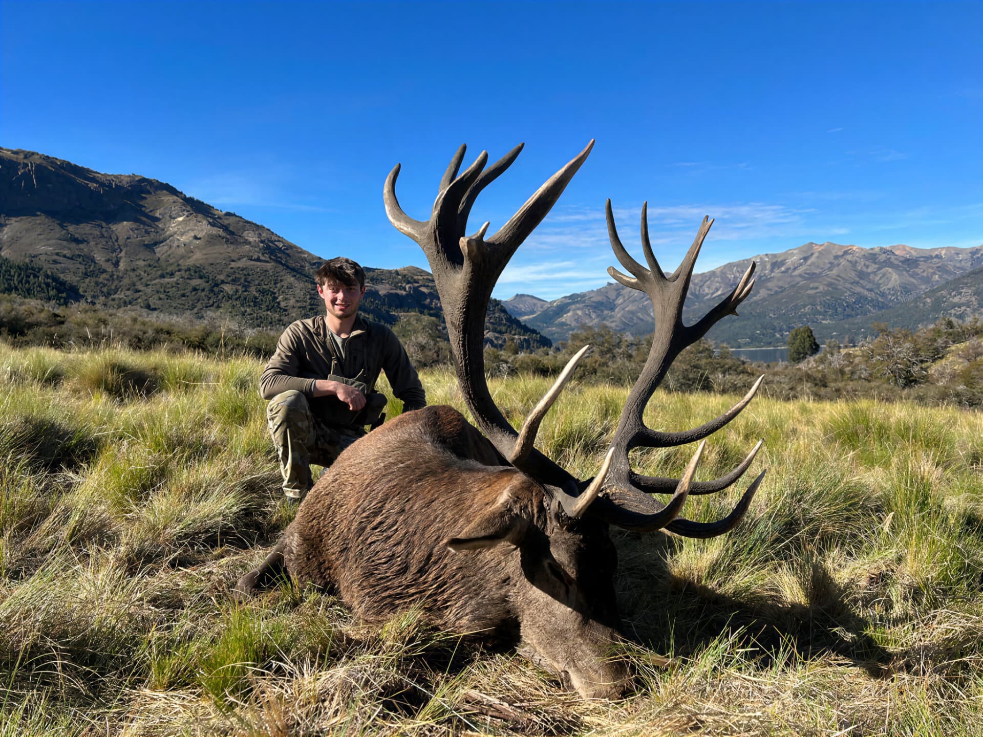 Hunter with a dark-antlered red stag on open grassland above the lake