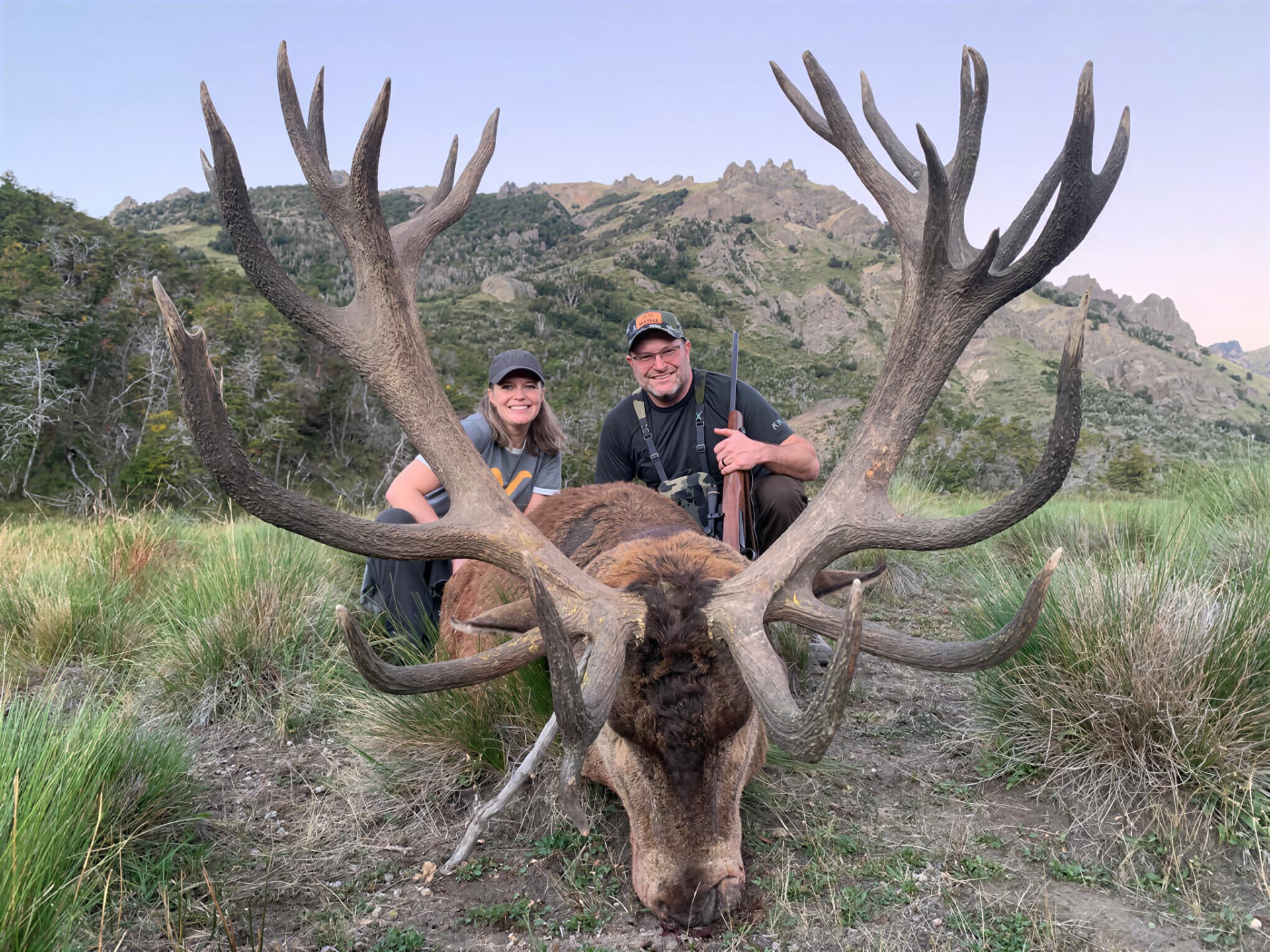 Couple behind a massive wide-racked red stag in a mountain meadow at dusk