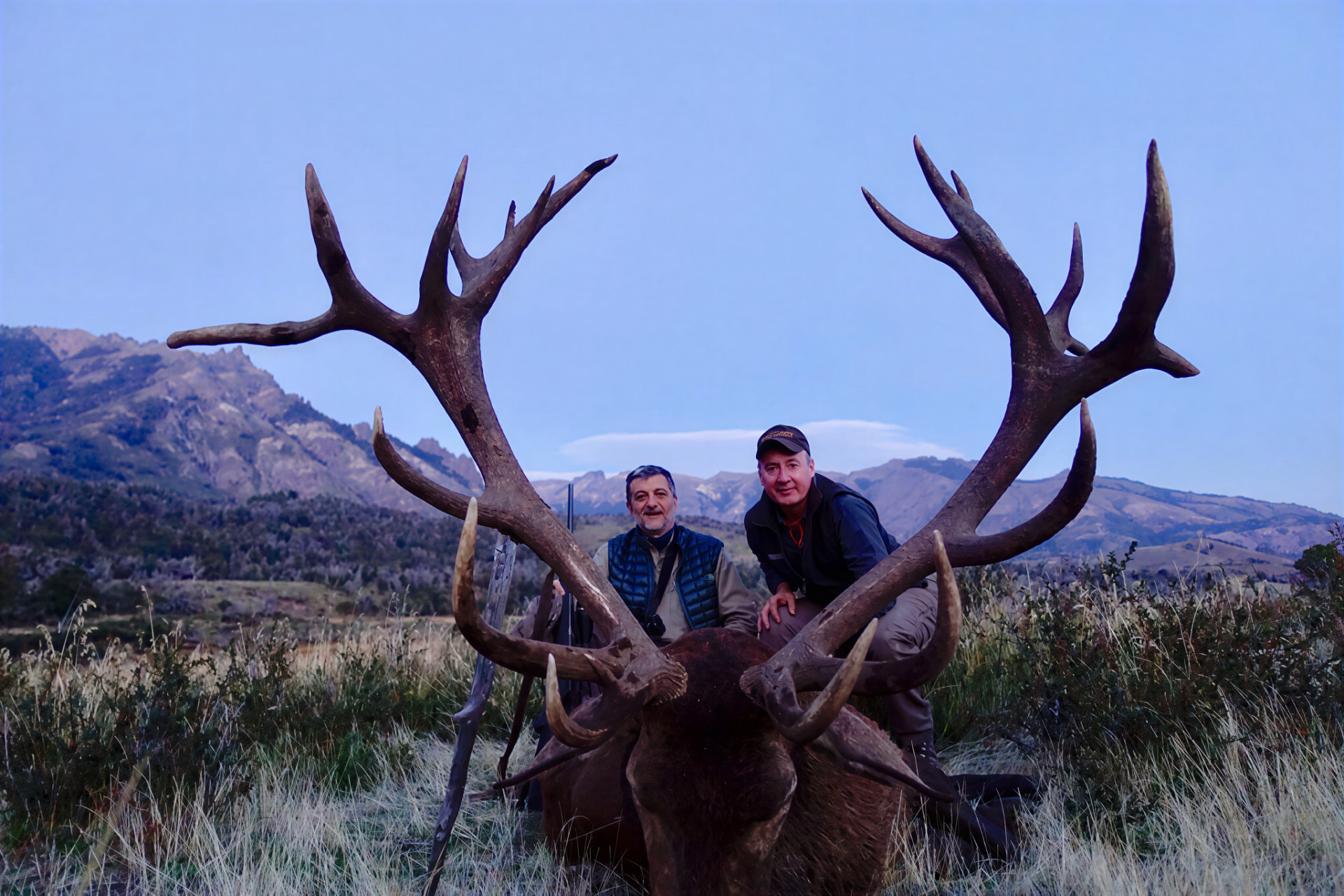 Two hunters with a giant red stag at dusk below the mountains