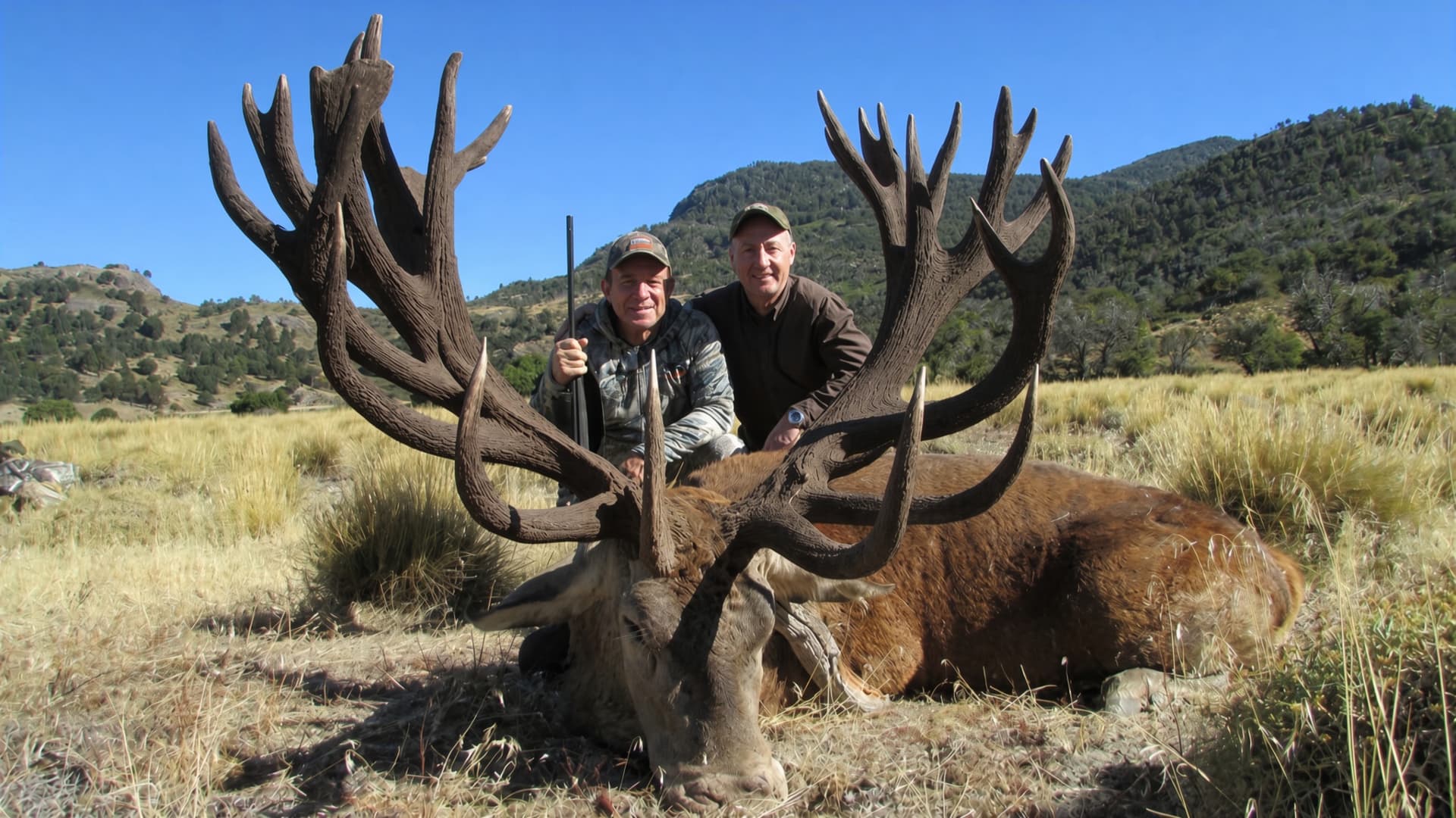 Two hunters behind a massive red stag with wide, palmated antlers in open Patagonian country