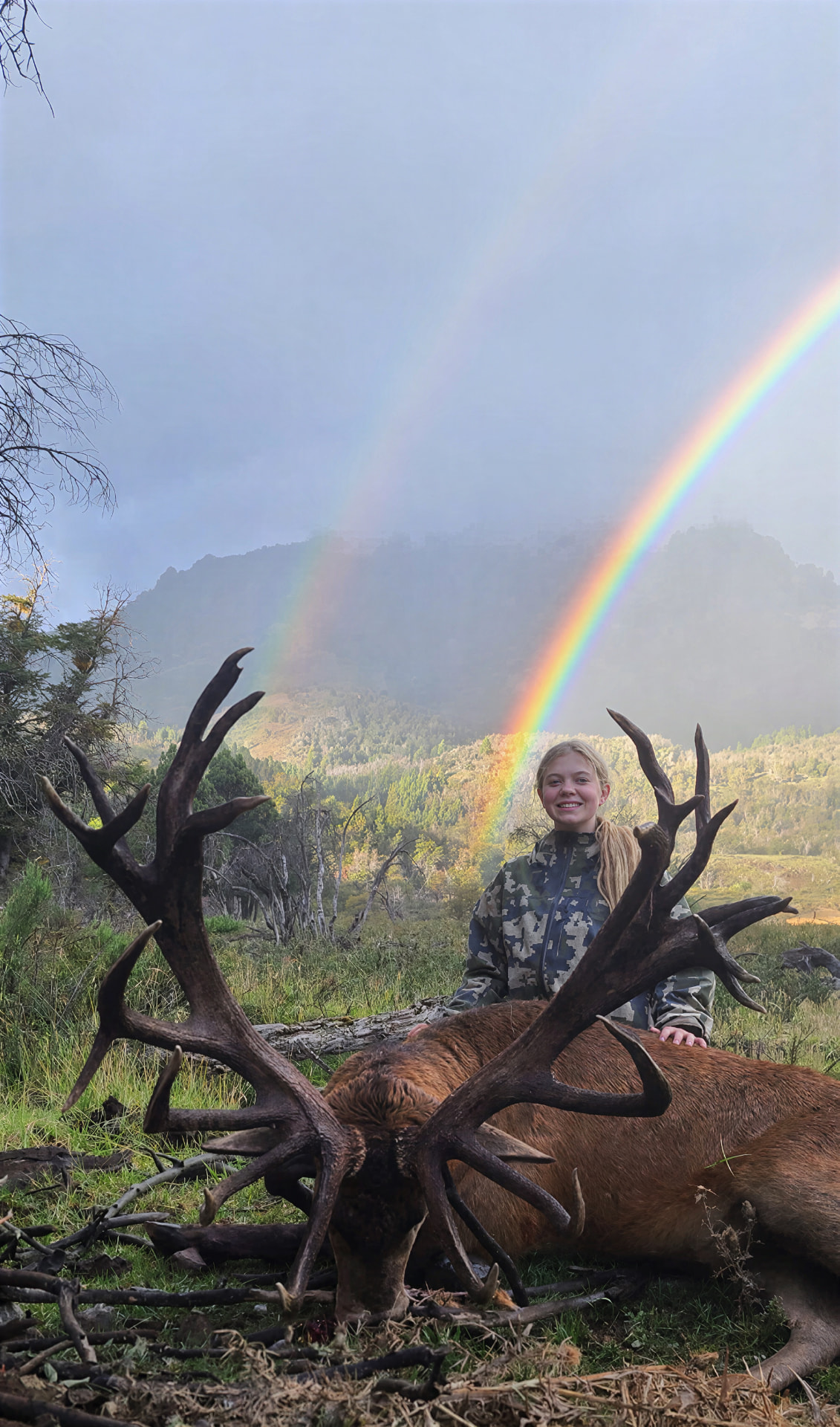 Young hunter with a red stag beneath a double rainbow in Patagonia