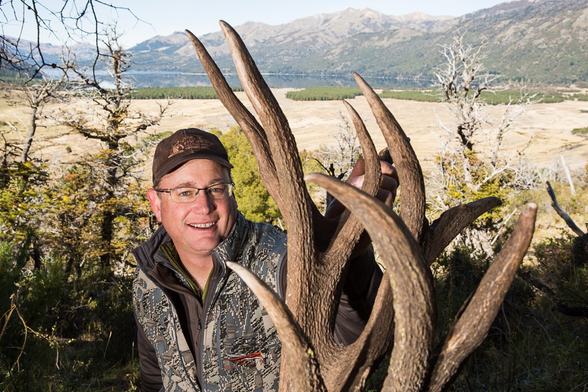 Smiling hunter behind the tall dark antlers of a Patagonian red stag above the lake