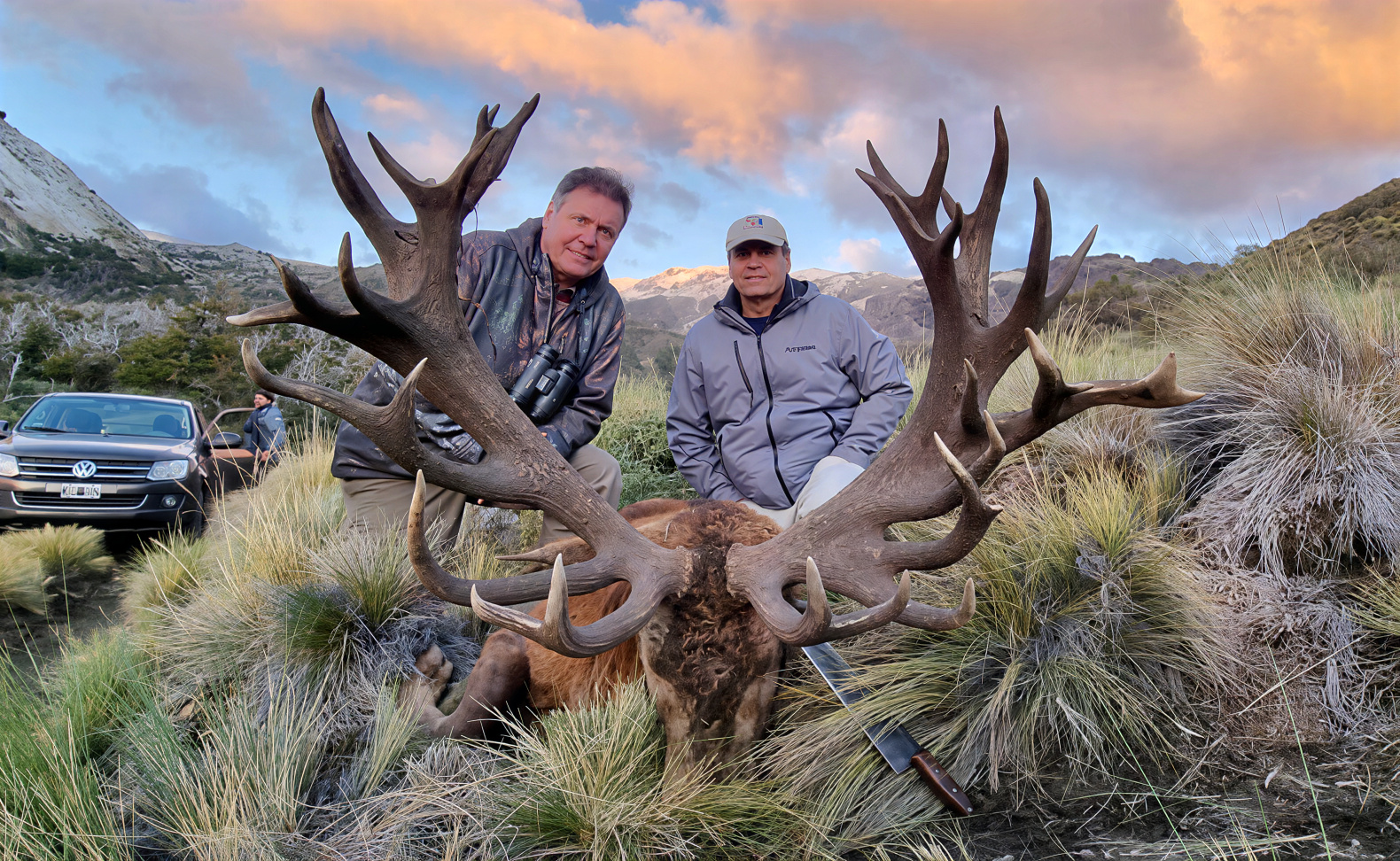 Two hunters behind a massive red stag with towering antlers at sunset in Patagonia