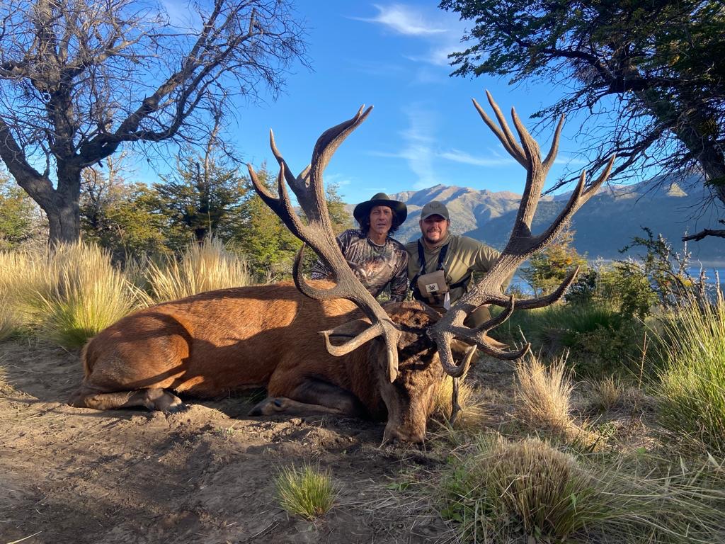 Two hunters with a heavy red stag beside a Patagonian lake