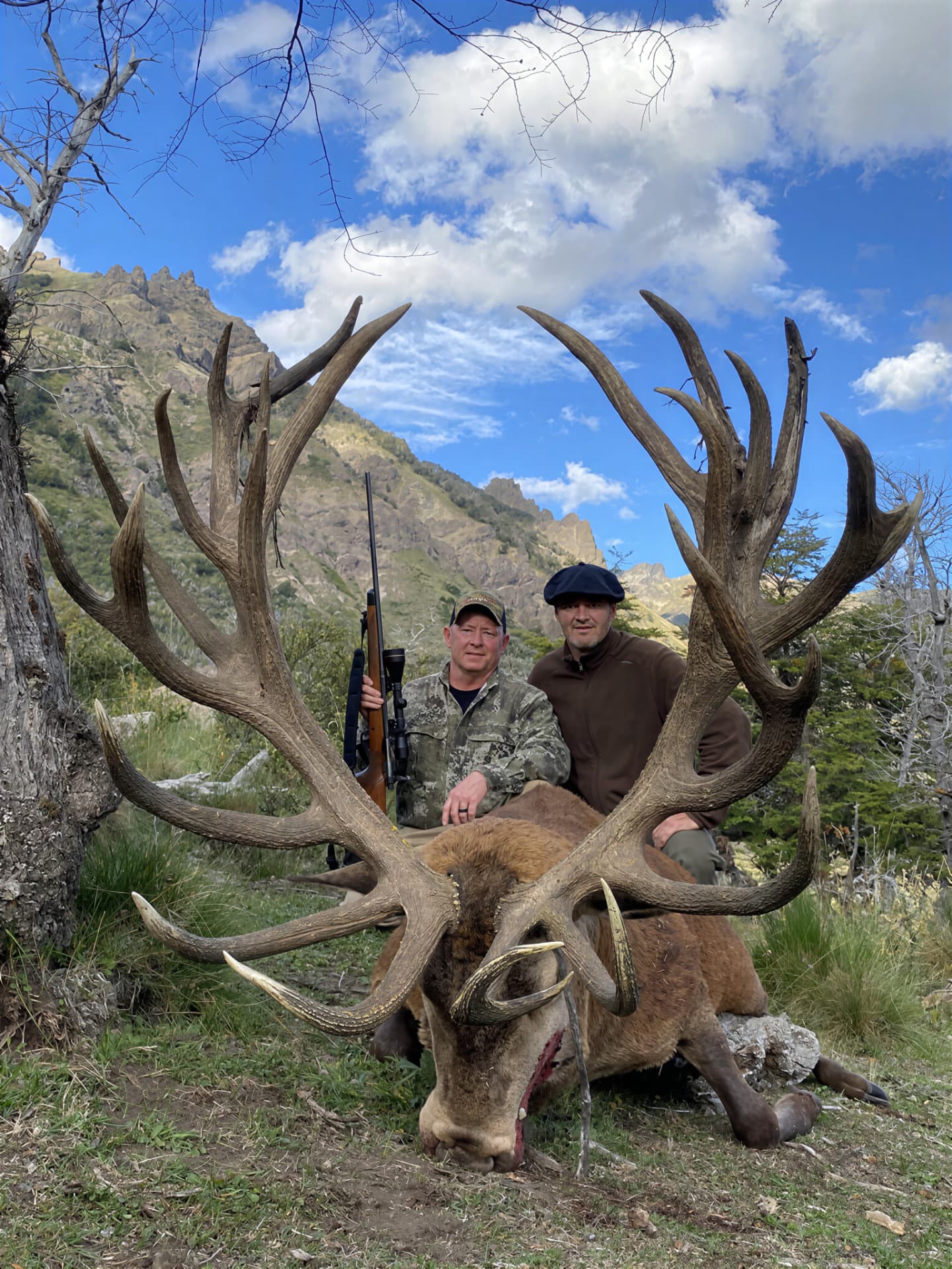 Hunter and guide with a gold-medal red stag below rocky Andean spires